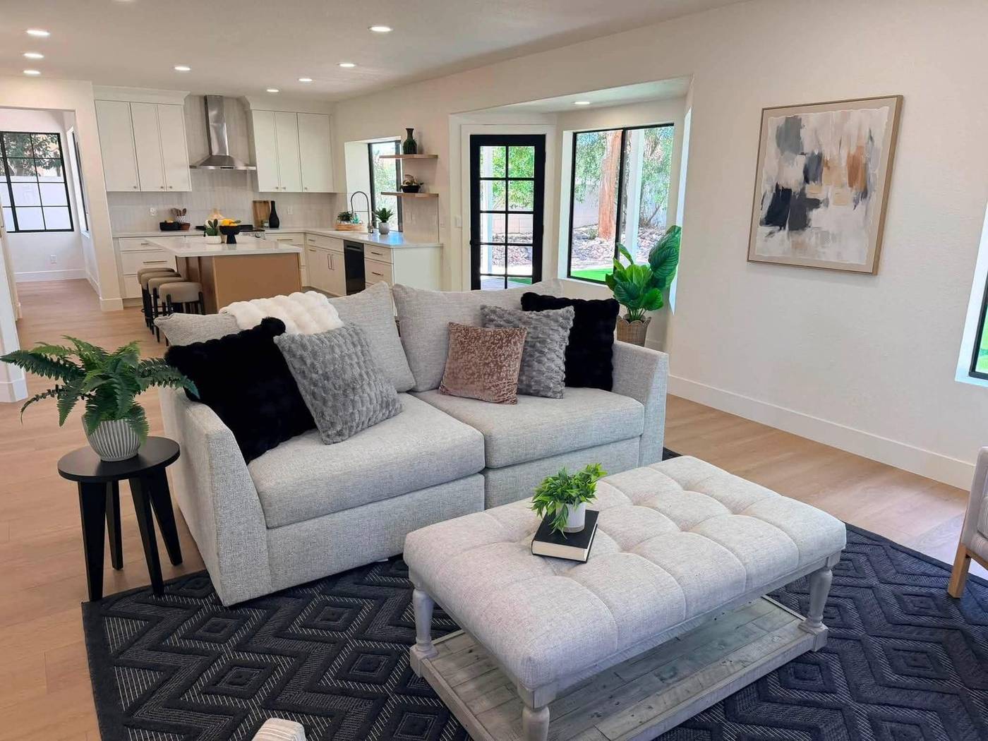 Mid-level staged living room with a gray sofa, tufted ottoman and open kitchen behind